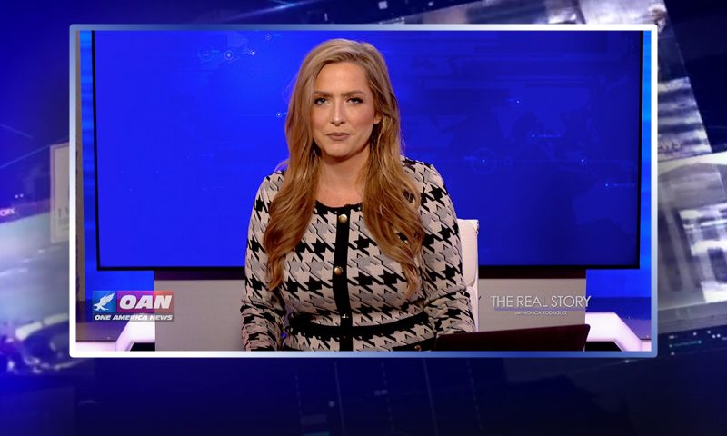 Video still of the host of The Real Story at the desk of their talk show on One America News Network.