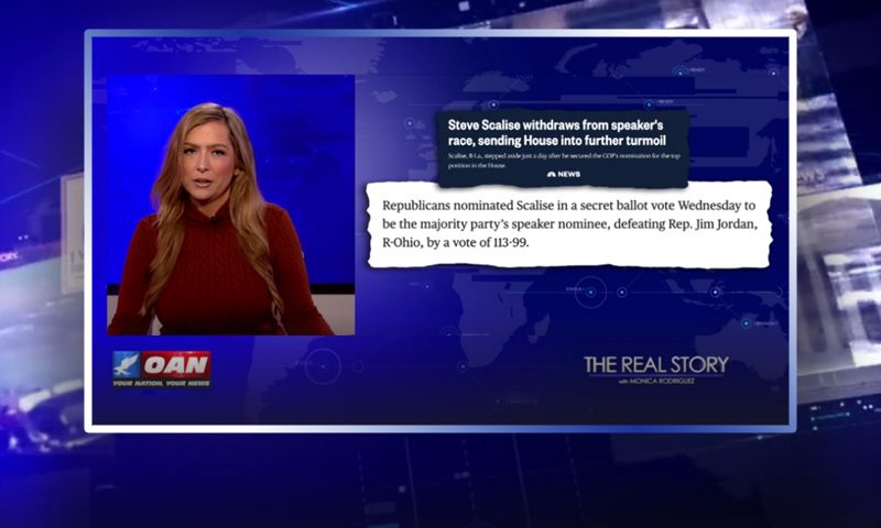 Video still of the host of The Real Story at the desk of their talk show on One America News Network.