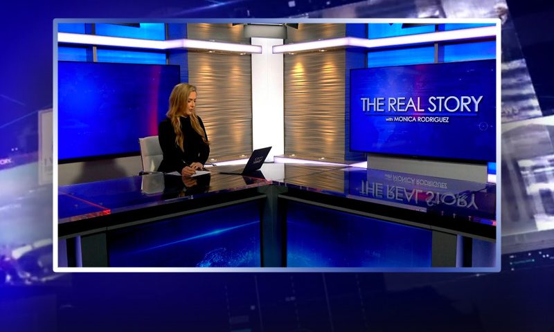 Video still of the host of The Real Story at the desk of their talk show on One America News Network.