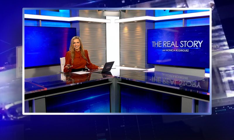Video still of the host of The Real Story at the desk of their talk show on One America News Network.