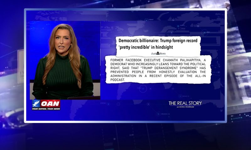 Video still of the host of The Real Story at the desk of their talk show on One America News Network.