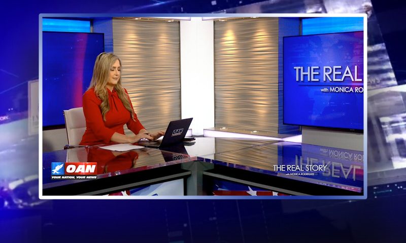 Video still of the host of The Real Story at the desk of their talk show on One America News Network.