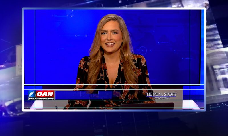 Video still of the host of The Real Story at the desk of their talk show on One America News Network.