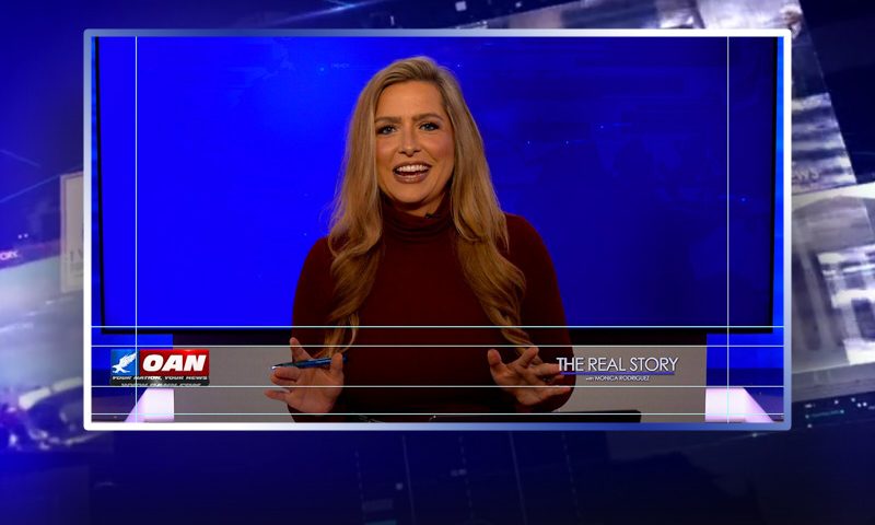 Video still of the host of The Real Story at the desk of their talk show on One America News Network.