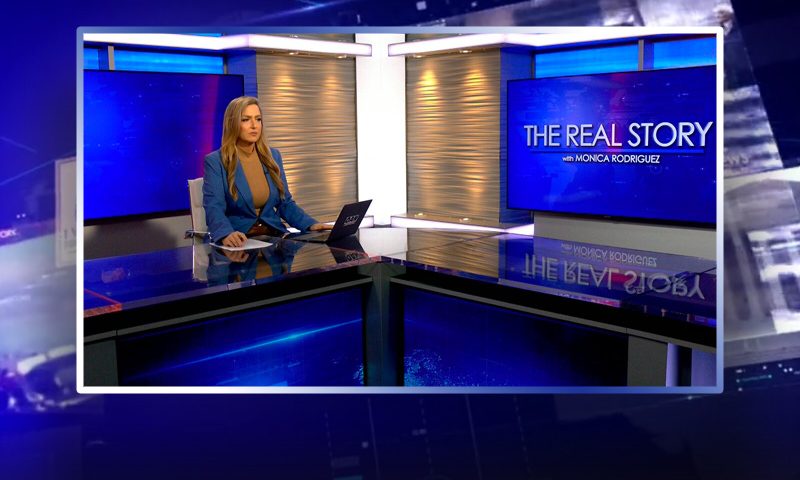 Video still of the host of The Real Story at the desk of their talk show on One America News Network.