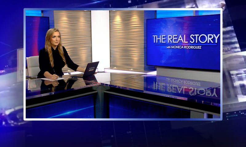 Video still of the host of The Real Story at the desk of their talk show on One America News Network.