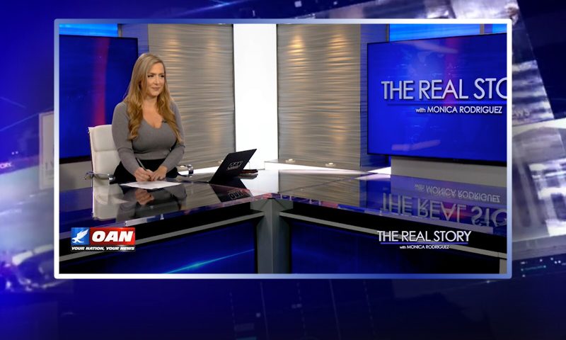 Video still of the host of The Real Story at the desk of their talk show on One America News Network.