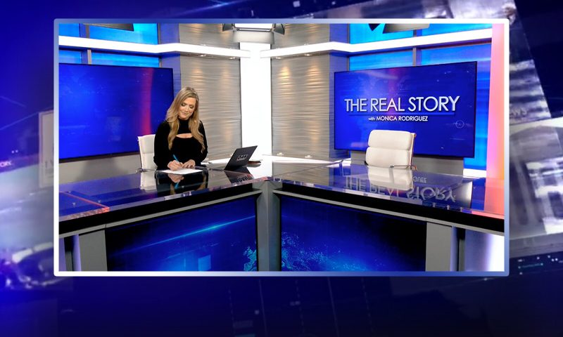 Video still of the host of The Real Story at the desk of their talk show on One America News Network.