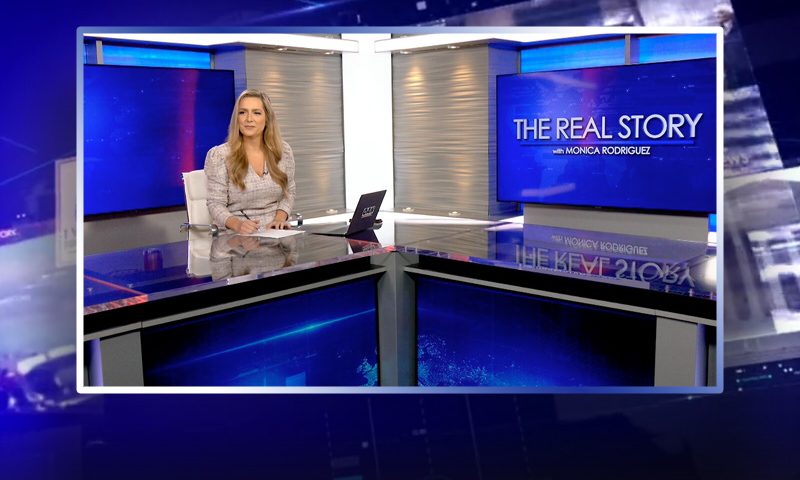 Video still of the host of The Real Story at the desk of their talk show on One America News Network.