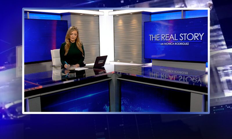 Video still of the host of The Real Story at the desk of their talk show on One America News Network.