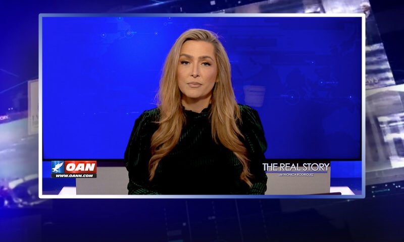 Video still of the host of The Real Story at the desk of their talk show on One America News Network.