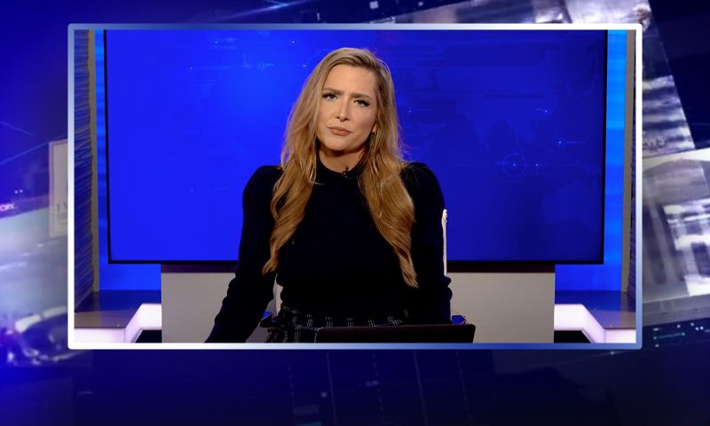 Video still of the host of The Real Story at the desk of their talk show on One America News Network.