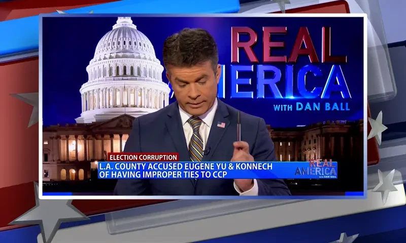 Video still of the host of Real America at the desk of their talk show on One America News Network.