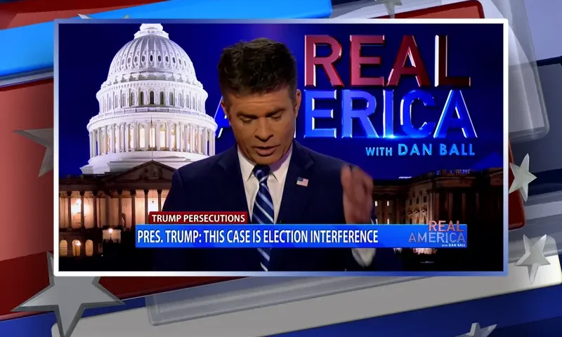 Video still of the host of Real America at the desk of their talk show on One America News Network.