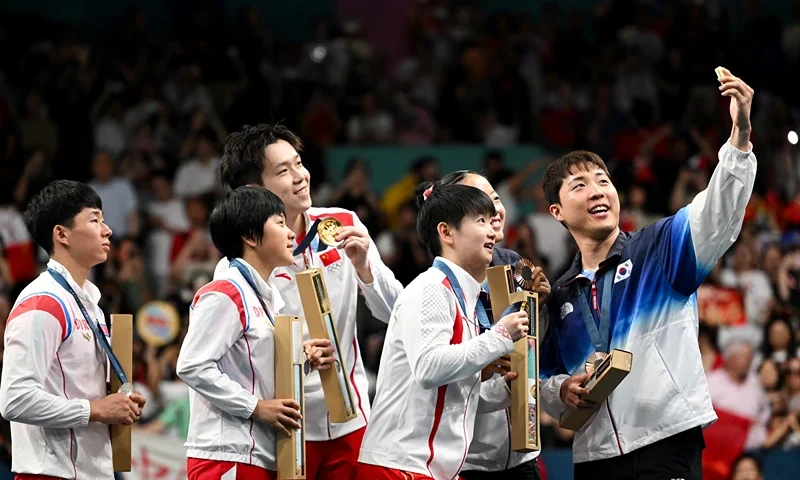TOPSHOT - Bronze medalists South Korea's Lim Jonghoon (R) takes a selfie picture with his mobile as he celebrates on the podium with : gold medalists China's Wang Chuqin (rear 2nd L) and China's Sun Yingsha (front 2nd L), silver medalists North Korea's Ri Jong Sik (L), and North Korea's Kim Kum Yong (front 2nd L) and bronze medallist and teammate South Korea's Shin Yubin (2nd R) at the end of their mixed table tennis doubles competition at the Paris 2024 Olympic Games at the South Paris Arena in Paris on July 30, 2024. (Photo by JUNG Yeon-je / AFP) (Photo by JUNG YEON-JE/AFP via Getty Images)