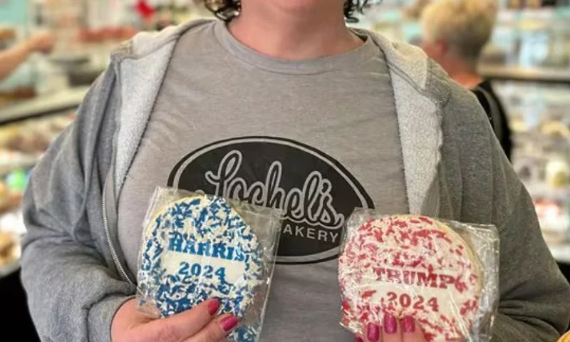 Bakery owner Kathleen Lochel holds sugar cookies, one with blue and white sprinkles and a Harris 2024 label on it, and the other, with red and white sprinkles and a Trump 2024 label on it at Lochel Bakery, on September 24, 2024, in Hatboro, a suburb of Philadelphia. (Photo via: Tassanee Vejpongsa/AP News)