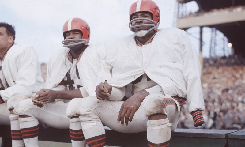 American football player Jim Brown sits on the bench during a game, wearing his helmet and uniform for the Cleveland Browns, circa 1960. (Photo by Hulton Archive/Getty Images)