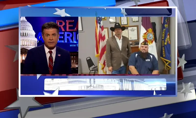 Video still from Real America on One America News Network showing a split screen of the host on the left side, and on the right side is the guest, Michael Carmichael.