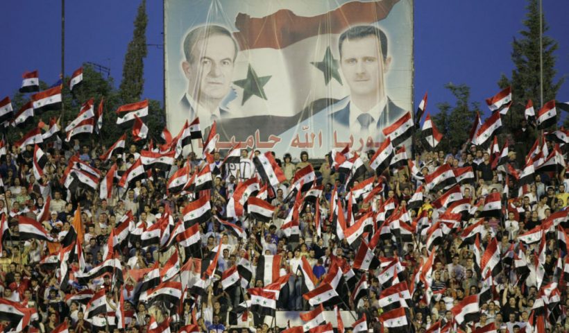 TOPSHOT - Syrian fans cheer for their national team under portraits of late president Hafez al-Assad and current President Bashar al-Assad (R) during their Asian World Cup qualifying football match against Kuwait at the Abbassin Stadium in Damascus on June 2, 2008. Syria won the match 1-0. AFP PHOTO/LOUAI BESHARA (Photo by Louai Beshara / AFP) (Photo by LOUAI BESHARA/AFP via Getty Images)