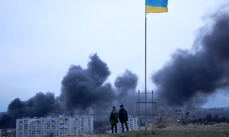 People standing near a Ukrainian national flag watch as dark smoke billows following an air strike in the western Ukrainian city of Lviv, on March 26, 2022. At least five people wounded in two strikes on Lviv, the regional governor said, in a rare attack on a city that has escaped serious fighting since Russian troops invaded last month. (Photo by Oleksii FILIPPOV / AFP) (Photo by OLEKSII FILIPPOV/AFP via Getty Images)