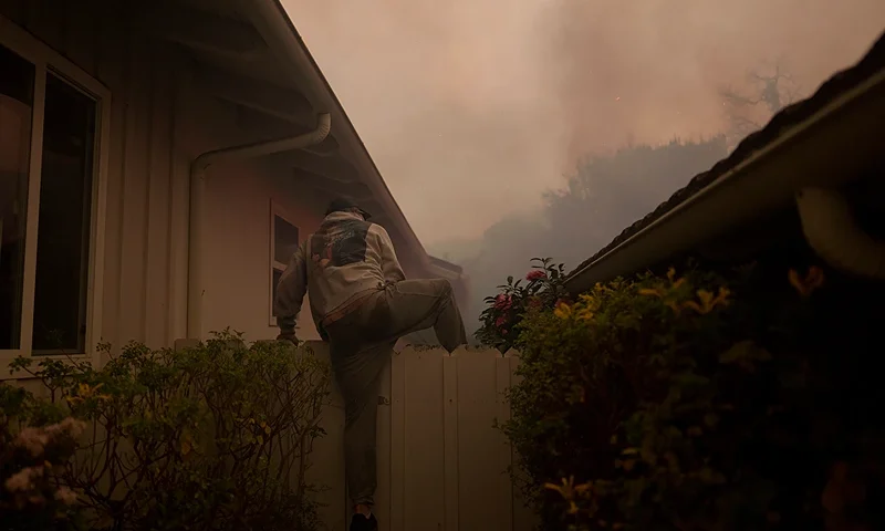 LOS ANGELES, CALIFORNIA - JANUARY 7: A resident climbs a fence to check on a home as flames from the Palisades Fire approach on January 7, 2025 in the Pacific Palisades neighborhood of Los Angeles, California. Fueled by intense Santa Ana Winds, the Palisades Fire has grown to over 2,900 acres and 30,000 people have been ordered to evacuate while a second fire has emerged near Eaton Canyon. (Photo by Eric Thayer/Getty Images)
