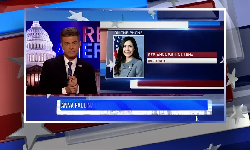 Video still of the host of Real America at the desk of their talk show on One America News Network.