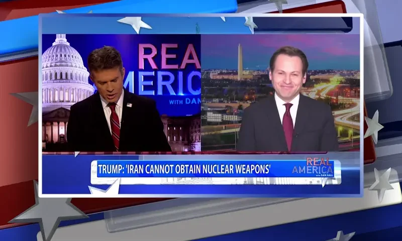 Video still from Real America on One America News Network showing a split screen of the host on the left side, and on the right side is the guest, Jacob Olidort.