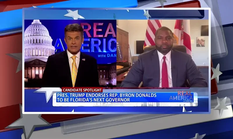Video still from Real America on One America News Network showing a split screen of the host on the left side, and on the right side is the guest, Byron Donalds.