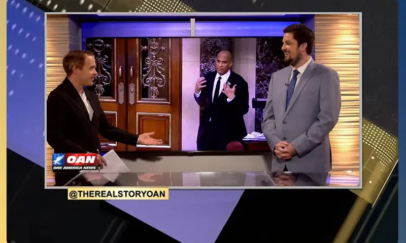 Video still of the host of The Real Story at the desk of their talk show on One America News Network.