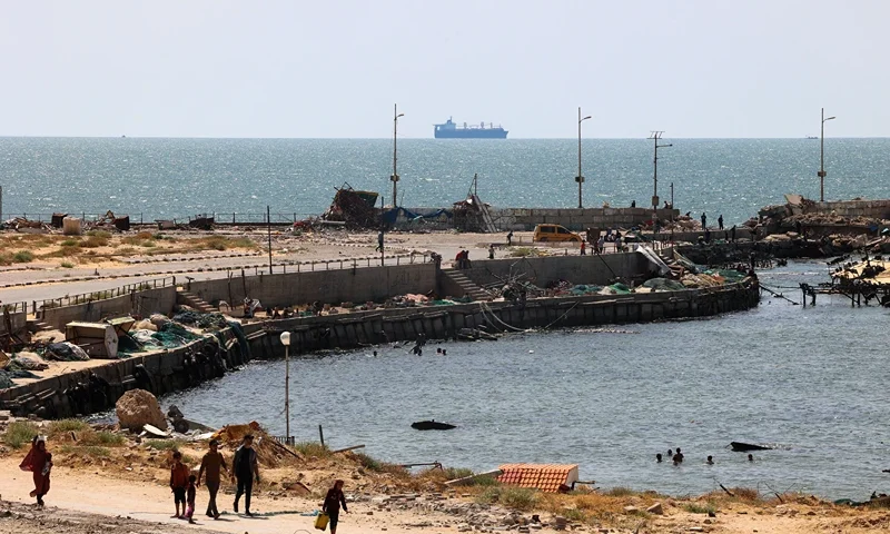 Palestinians walk past a jetty in Gaza City with a view of navy vessels off the coast as part of a humanitarian "maritime corridor" announced by US Central Command (CENTCOM) on May 17, 2024 amid the ongoing conflict in the Gaza Strip between Israel and Hamas. The US military said aid deliveries began on May 17 via a temporary pier in Gaza aimed at ramping up emergency humanitarian assistance to the war-ravaged Palestinian territory. (Photo by AFP) (Photo by -/AFP via Getty Images)