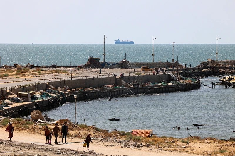 Palestinians walk past a jetty in Gaza City with a view of navy vessels off the coast as part of a humanitarian "maritime corridor" announced by US Central Command (CENTCOM) on May 17, 2024 amid the ongoing conflict in the Gaza Strip between Israel and Hamas. The US military said aid deliveries began on May 17 via a temporary pier in Gaza aimed at ramping up emergency humanitarian assistance to the war-ravaged Palestinian territory. (Photo by AFP) (Photo by -/AFP via Getty Images)