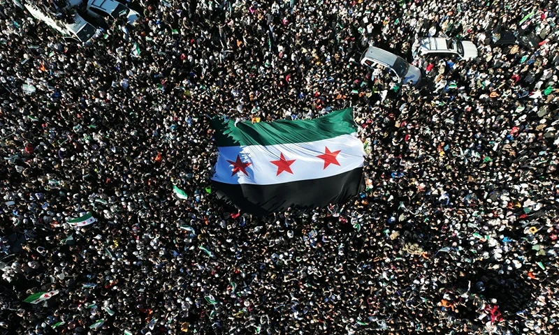 TOPSHOT - An aerial photo shows crowds of Syrians raising a giant independence-era flag, used by the opposition since the uprising began in 2011, as they celebrate the fall of Bashar al-Assad's iron-fisted rule earlier this week at the central Umayyad Square in Damascus on December 13, 2024. Thousands of jubilant Syrians also gathered outside a landmark mosque in the capital Damascus to celebrate during the first Friday prayers since Assad's ouster on December 8. (Photo by Omar HAJ KADOUR / AFP) (Photo by OMAR HAJ KADOUR/AFP via Getty Images)