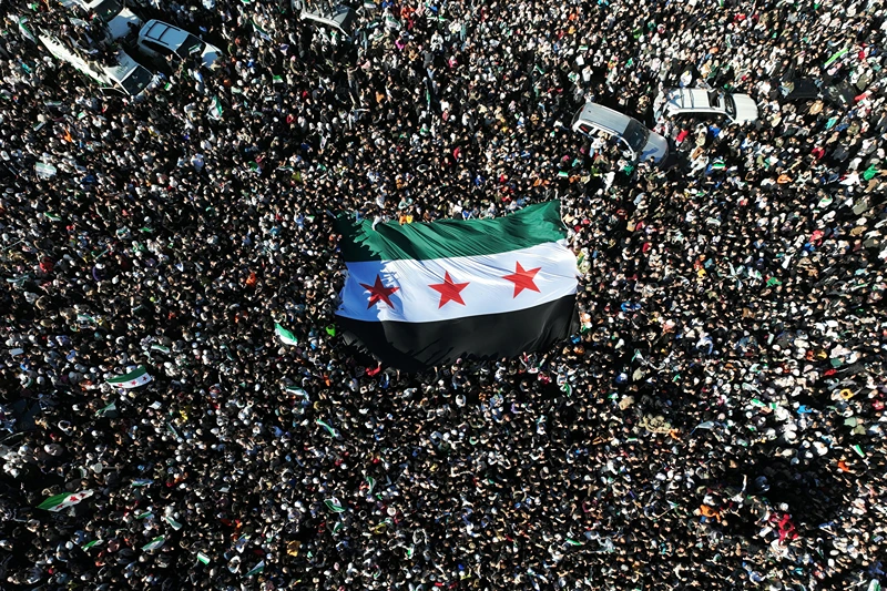TOPSHOT - An aerial photo shows crowds of Syrians raising a giant independence-era flag, used by the opposition since the uprising began in 2011, as they celebrate the fall of Bashar al-Assad's iron-fisted rule earlier this week at the central Umayyad Square in Damascus on December 13, 2024. Thousands of jubilant Syrians also gathered outside a landmark mosque in the capital Damascus to celebrate during the first Friday prayers since Assad's ouster on December 8. (Photo by Omar HAJ KADOUR / AFP) (Photo by OMAR HAJ KADOUR/AFP via Getty Images)