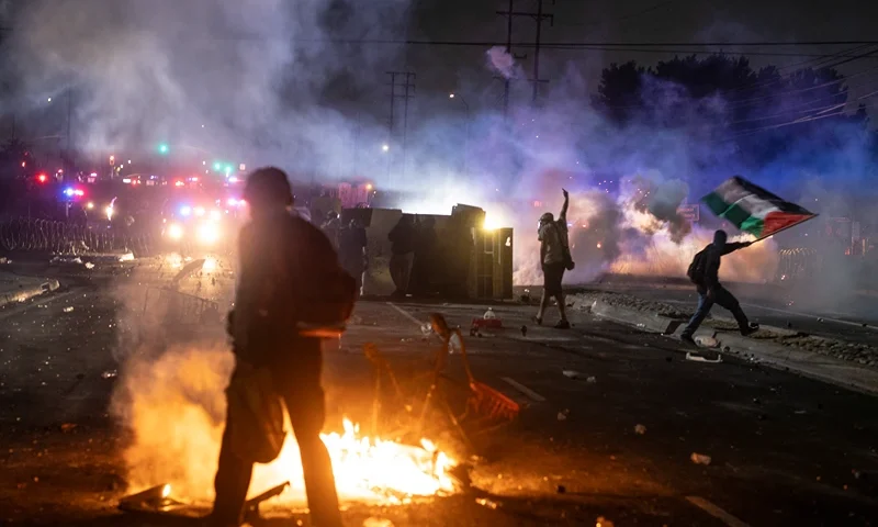 A protester waves the Palestinian flag (R) as law enforcement clashes with demonstrators during a protest following federal immigration operations, in the Compton neighborhood of Los Angeles, California on June 7, 2025. US President Donald Trump deployed 2,000 troops on June 7, 2025 to handle escalating protests against immigration enforcement raids in the Los Angeles area, a move the state's governor termed "purposefully inflammatory." Federal agents clashed with angry crowds in a Los Angeles suburb as protests stretched into a second night Saturday, shooting flash-bang grenades and shutting part of a freeway amid raids on undocumented migrants, reports said. (Photo by RINGO CHIU / AFP) (Photo by RINGO CHIU/AFP via Getty Images)