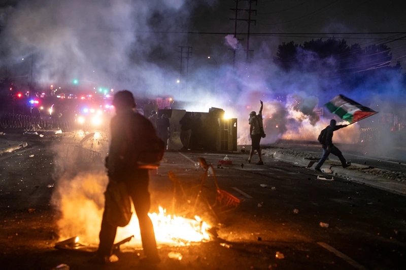A protester waves the Palestinian flag (R) as law enforcement clashes with demonstrators during a protest following federal immigration operations, in the Compton neighborhood of Los Angeles, California on June 7, 2025. US President Donald Trump deployed 2,000 troops on June 7, 2025 to handle escalating protests against immigration enforcement raids in the Los Angeles area, a move the state's governor termed "purposefully inflammatory." Federal agents clashed with angry crowds in a Los Angeles suburb as protests stretched into a second night Saturday, shooting flash-bang grenades and shutting part of a freeway amid raids on undocumented migrants, reports said. (Photo by RINGO CHIU / AFP) (Photo by RINGO CHIU/AFP via Getty Images)