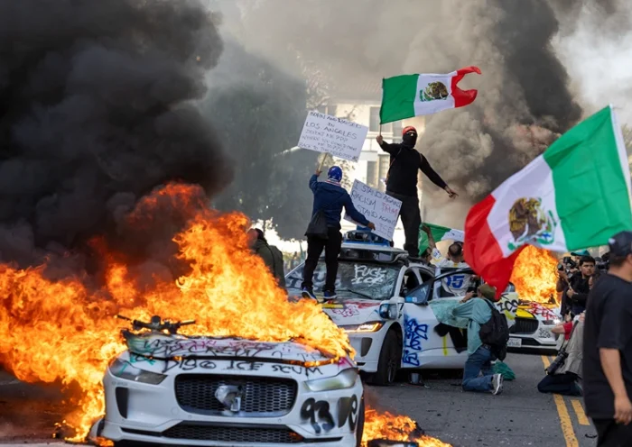Demonstrators wave flags from atop a wrecked car, standing beside burning Waymo vehicles, as protesters clash with law enforcement in the streets surrounding the federal building during a protest following federal immigration operations in Los Angeles, California, on June 8, 2025. Demonstrators torched cars and scuffled with security forces in Los Angeles on June 8, as police kept protestors away from the National Guard troops President Donald Trump sent to the streets of the second biggest US city. (Photo by RINGO CHIU / AFP) (Photo by RINGO CHIU/AFP via Getty Images)