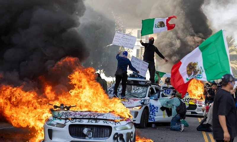 Demonstrators wave flags from atop a wrecked car, standing beside burning Waymo vehicles, as protesters clash with law enforcement in the streets surrounding the federal building during a protest following federal immigration operations in Los Angeles, California, on June 8, 2025. Demonstrators torched cars and scuffled with security forces in Los Angeles on June 8, as police kept protestors away from the National Guard troops President Donald Trump sent to the streets of the second biggest US city. (Photo by RINGO CHIU / AFP) (Photo by RINGO CHIU/AFP via Getty Images)