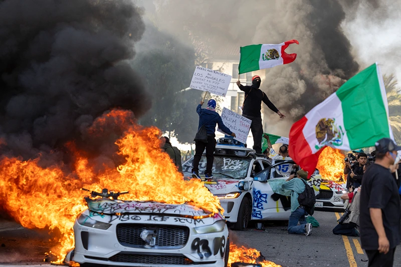 Demonstrators wave flags from atop a wrecked car, standing beside burning Waymo vehicles, as protesters clash with law enforcement in the streets surrounding the federal building during a protest following federal immigration operations in Los Angeles, California, on June 8, 2025. Demonstrators torched cars and scuffled with security forces in Los Angeles on June 8, as police kept protestors away from the National Guard troops President Donald Trump sent to the streets of the second biggest US city. (Photo by RINGO CHIU / AFP) (Photo by RINGO CHIU/AFP via Getty Images)