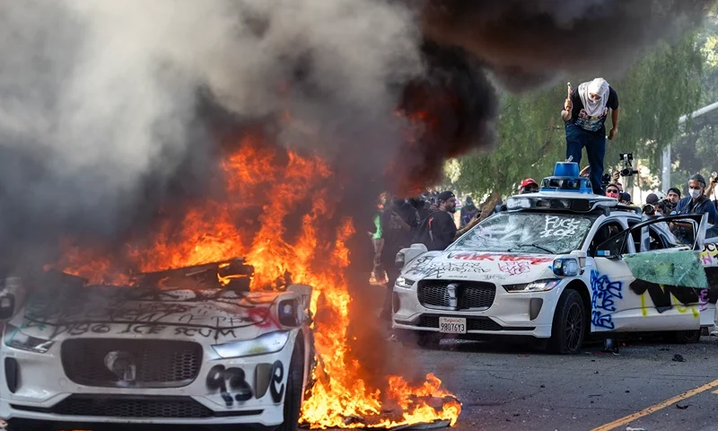 Demonstrators smash the windshield of a vehicle next to a burning Waymo vehicle as protesters clash with law enforcement in the streets surrounding the federal building during a protest following federal immigration operations in Los Angeles, California, on June 8, 2025. Demonstrators torched cars and scuffled with security forces in Los Angeles on June 8, as police kept protestors away from the National Guard troops President Donald Trump sent to the streets of the second biggest US city. (Photo by RINGO CHIU / AFP) (Photo by RINGO CHIU/AFP via Getty Images)