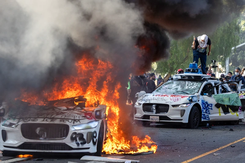 Demonstrators smash the windshield of a vehicle next to a burning Waymo vehicle as protesters clash with law enforcement in the streets surrounding the federal building during a protest following federal immigration operations in Los Angeles, California, on June 8, 2025. Demonstrators torched cars and scuffled with security forces in Los Angeles on June 8, as police kept protestors away from the National Guard troops President Donald Trump sent to the streets of the second biggest US city. (Photo by RINGO CHIU / AFP) (Photo by RINGO CHIU/AFP via Getty Images)