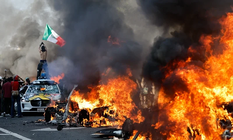 LOS ANGELES, LOS ANGELES - JUNE 08: A protestor holds up a Mexican flag as burning cars line the street on June 08, 2025 in Los Angeles, California. Tensions in the city remain high after the Trump administration called in the National Guard against the wishes of city leaders following two days of clashes with police during a series of immigration raids. (Photo by Mario Tama/Getty Images)