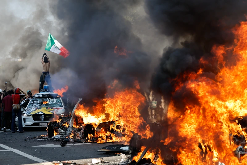 LOS ANGELES, LOS ANGELES - JUNE 08: A protestor holds up a Mexican flag as burning cars line the street on June 08, 2025 in Los Angeles, California. Tensions in the city remain high after the Trump administration called in the National Guard against the wishes of city leaders following two days of clashes with police during a series of immigration raids. (Photo by Mario Tama/Getty Images)