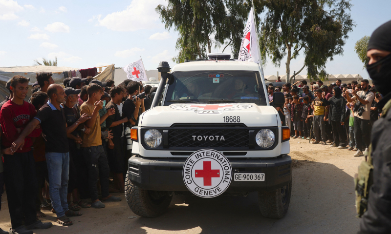 A Qassam Brigades militant stands guard as a crowd of Palestinians watches a vehicle of the International Committee of the Red Cross (ICRC) carrying Israeli hostages leave the area south of Deir el-Balah in the central Gaza Strip to handover the released Israelis on October 13, 2025. Israel said that the last 20 living hostages released by Hamas on October 13 had arrived in the country. (Photo by Bashar TALEB / AFP) (Photo by BASHAR TALEB/AFP via Getty Images)