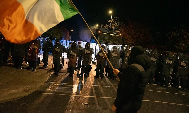 DUBLIN, IRELAND - OCTOBER 21: A protester waves the Irish flag in front of riot police on October 21, 2025 in Dublin, Ireland. Protesters have gathered outside the Citywest Hotel in Dublin after police arrested a foreign national male on Sunday for the rape of a 10-year-old girl. (Photo by Natalia Campos/Getty Images)