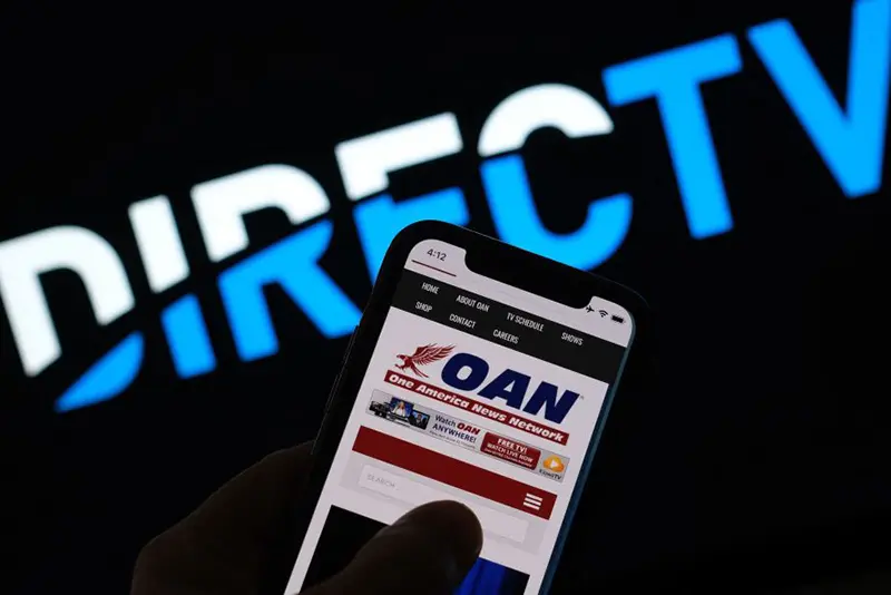 This illustration photo shows a person reading the One America Network website on a smartphone in front of a DirecTV logo. (Photo by Chris DELMAS / AFP) (Photo by CHRIS DELMAS/AFP via Getty Images)