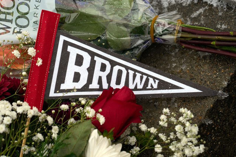 A Brown University pennant is covered by flowers left by mourners at a makeshift memorial outside the Barus & Holley engineering building on the campus of Brown University, in Providence, Rhode Island on December 14, 2025. US authorities on Sunday detained a person of interest in the mass shooting at Brown University that left two people dead and nine others wounded, the latest in a long line of school attacks nationwide. (Photo by Bing Guan / AFP via Getty Images)