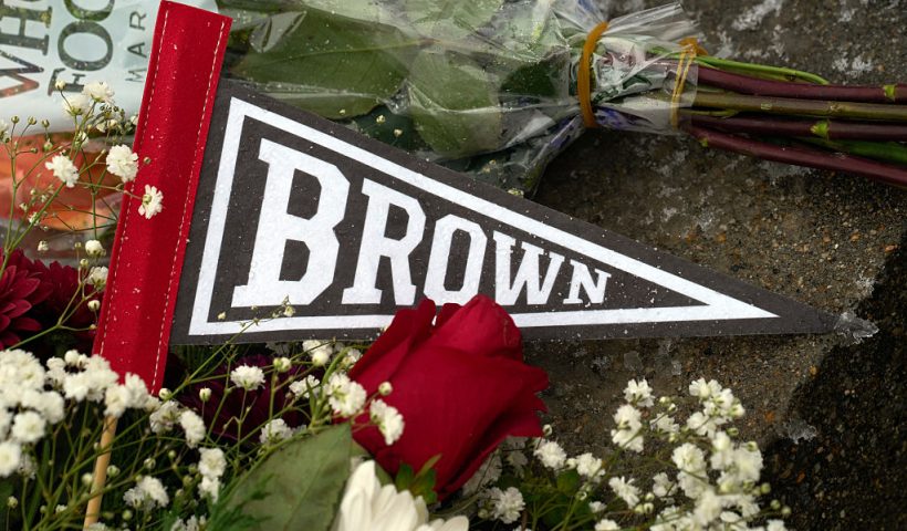 A Brown University pennant is covered by flowers left by mourners at a makeshift memorial outside the Barus & Holley engineering building on the campus of Brown University, in Providence, Rhode Island on December 14, 2025. US authorities on Sunday detained a person of interest in the mass shooting at Brown University that left two people dead and nine others wounded, the latest in a long line of school attacks nationwide. (Photo by Bing Guan / AFP via Getty Images)