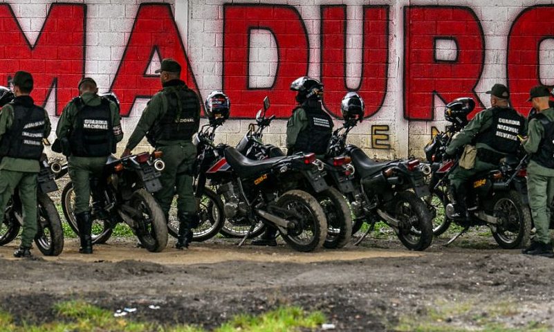 Members of the National Guard stand guard in front of a mural of ousted Venezuela's President Nicolas Maduro in Caracas on January 10, 2026. The US State Department on January 10, urged Americans in Venezuela to leave the country "immediately," citing risks from armed militias searching vehicles for US citizens at roadblocks. (Photo by Juan BARRETO / AFP via Getty Images)
